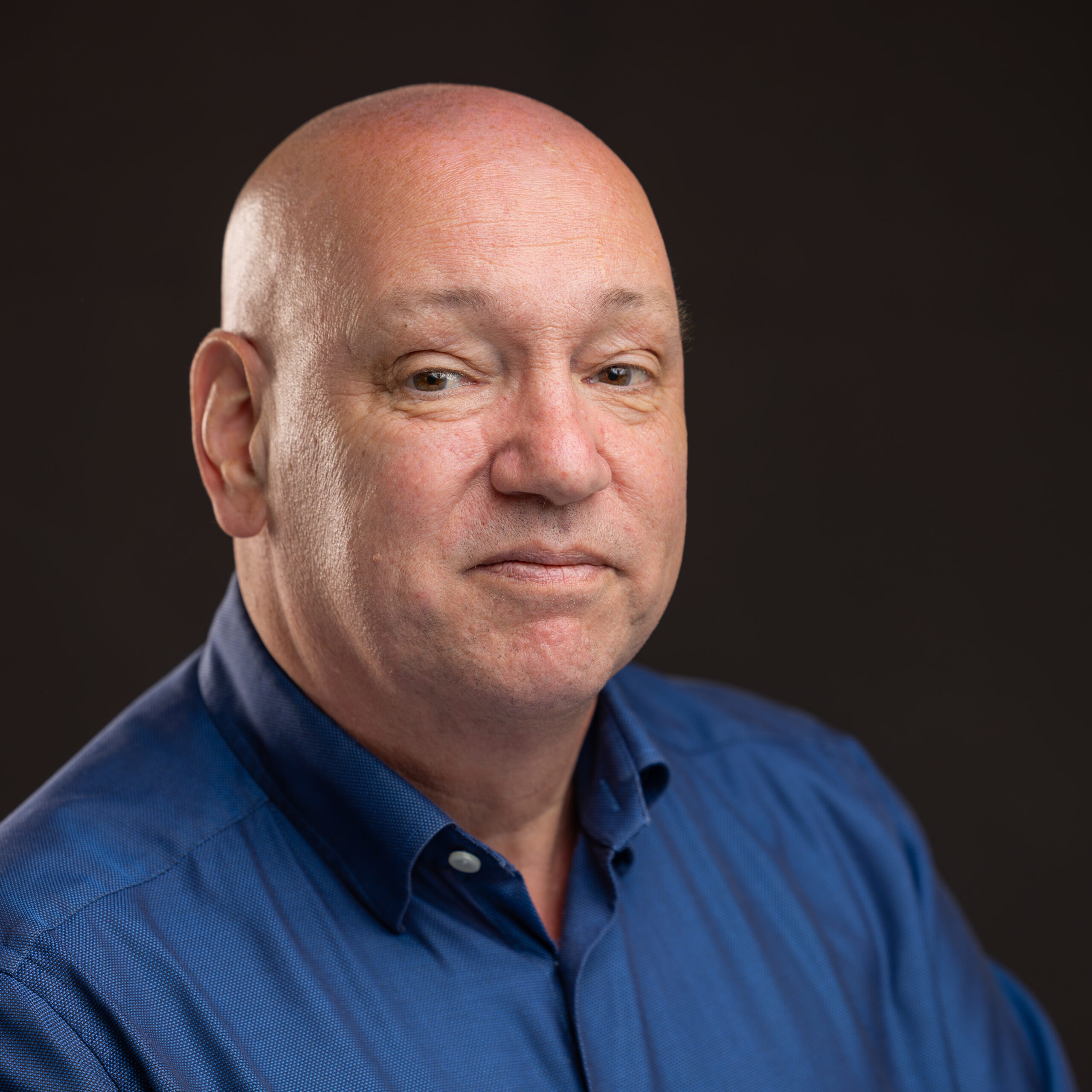 A middle-aged bald man wearing a blue button-up shirt poses in front of a dark background, looking at the camera with a neutral expression.