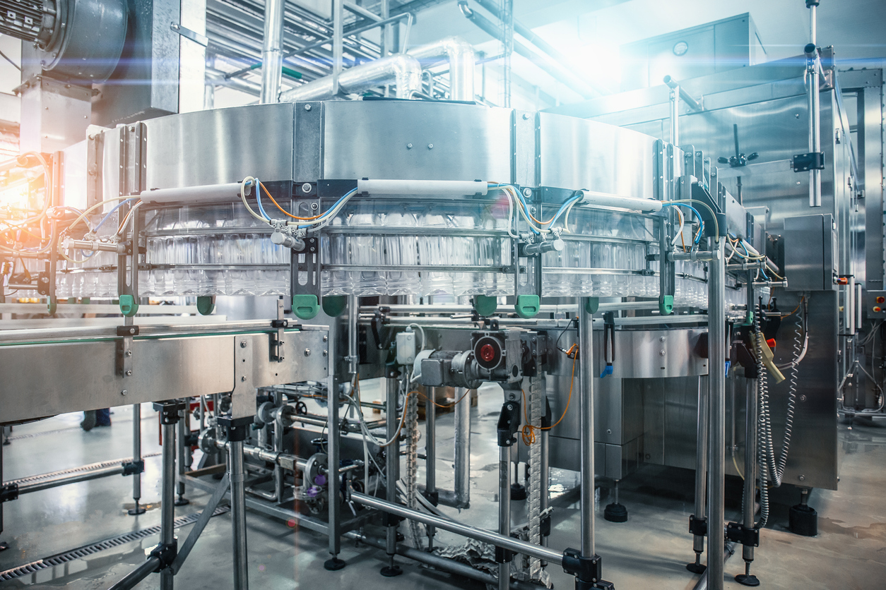 Een moderne geautomatiseerde bottelarij met doorzichtige plastic flessen die over een transportband bewegen, omringd door roestvrijstalen machines en industriële pijpen in een helder verlichte, schone omgeving.